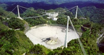 Photograph of a radio telescope dish set into the landscape, surrounded by towers that support suspended cables across its face.