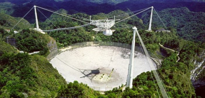 Photograph of a radio telescope dish set into the landscape, surrounded by towers that support suspended cables across its face.