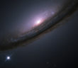 A spiral galaxy seen nearly edge-on. The galaxy has a bright center that is surrounded by a disk of dark black and brown clouds. Dim blue light can be seen through the clouds. To the lower left of the galaxy, just below the disk, is a bright white object with diffraction spikes. The background is mostly dark, scattered with a few small dim white objects.