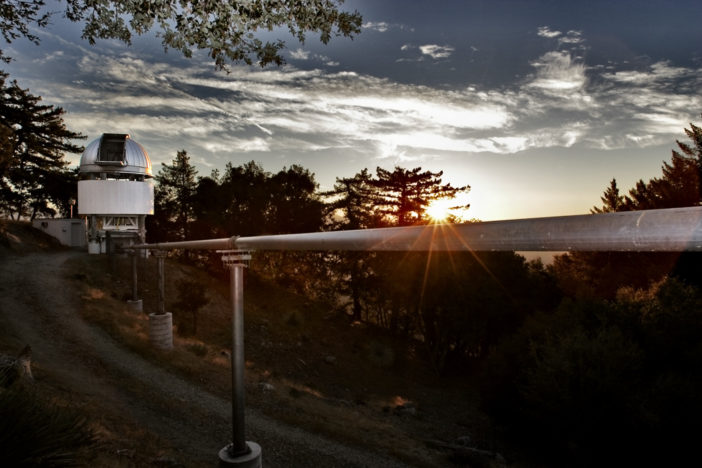 An open telescope dome is seen at the left of the image, with a pipe leaving it on the right. The pipe stretches across the entire image, moving into the foreground. The rising sun is roughly in the center of the image. The background is moderately populated with trees.