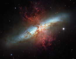 A cigar-shaped band of soft, diffuse light from the plane of M82 is crisscrossed by dark dust clouds.