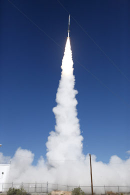 A long, skinny rocket launched into space, trailed by puffy white exhaust clouds. In the foreground, there is a chain-link fence, a telephone pole, and some scraggly bushes.
