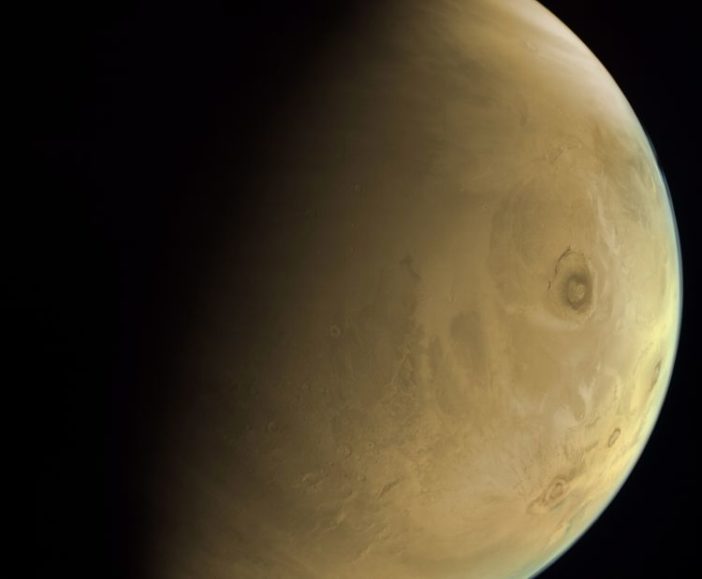 A photograph of Mars, half in shadow. Olympus Mons, a huge shield volcano, sits off-center on Mars's disk.