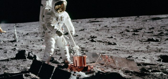 A photograph of an astronaut on the lunar surface standing over an instrument with large solar panels