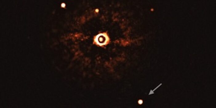 A photograph of a masked-out star surrounded by a halo of light leaking around the edge of the mask. Surrounding the halo are smaller star-like dots, one of which is annotated with an arrow. This is the directly imaged planet, and the other dots are faint background stars.