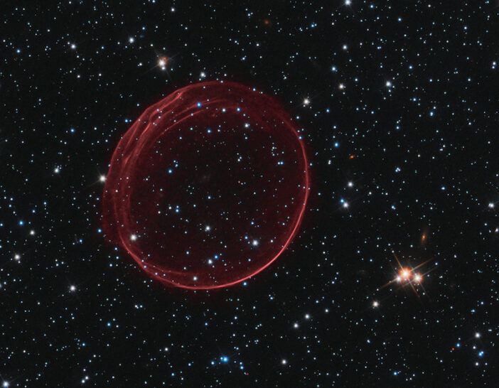 A circular red gas bubble, more opaque on the edges than in the center, sits in a field of stars.