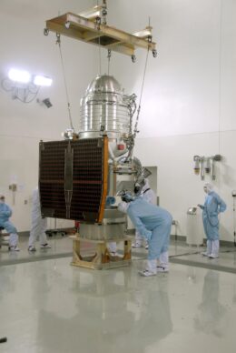 A photograph of a large silver cylinder sitting vertically within a clean room. It is surrounded by four technicians in protective clothing. They are roughly half the height of the telescope.