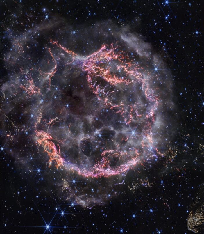 Cassiopeia A, a circular-shaped cloud of gas and dust with complex structure. The inner shell is made of bright pink and orange filaments studded with clumps and knots that look like tiny pieces of shattered glass. Around the exterior of the inner shell, particularly at the upper right, there are curtains of wispy gas that look like campfire smoke. The white smoke-like material also appears to fill the cavity of the inner shell, featuring structures shaped like large bubbles. Around and within the nebula, there are various stars seen as points of blue and white light. Outside the nebula, there are also clumps of yellow dust, with a particularly large clump at the bottom right corner that appears to have very detailed striations.