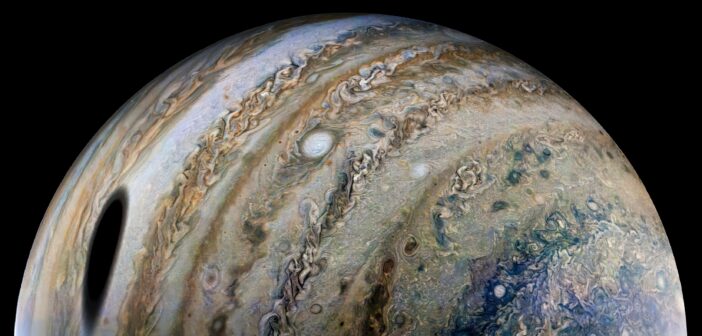 A close-up photograph of Jupiter. Dramatic cloud bands encircle the planet, and a large dark shadow of a nearby moon appears at left.