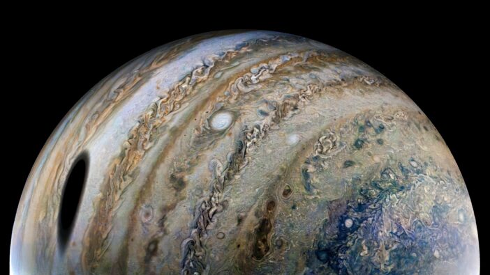 A close-up photograph of Jupiter. Dramatic cloud bands encircle the planet, and a large dark shadow of a nearby moon appears at left.